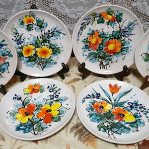 Może przedstawiać: Zestaw sześciu białych ceramicznych talerzy obiadowych z kwiatowym wzorem. Talerze zdobią różne kolorowe kwiaty, w tym żółte, pomarańczowe i czerwone, na białym tle. Kwiaty są malowane w stylizowany, abstrakcyjny sposób.