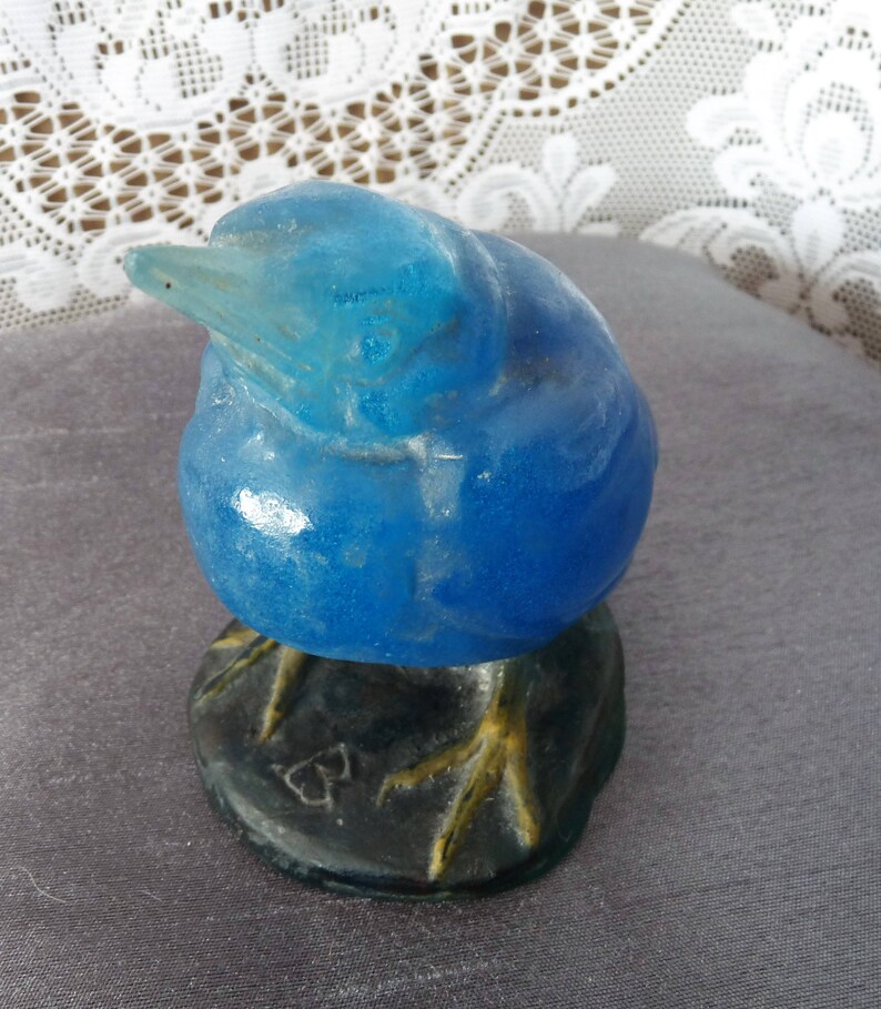 Pode incluir: Uma figurinha de p&aacute;ssaro de vidro azul com p&eacute;s amarelos e uma base preta. O p&aacute;ssaro est&aacute; empoleirado em uma rocha com um pequeno desenho em forma de cora&ccedil;&atilde;o.