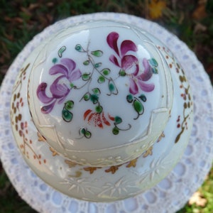 May include: A white porcelain lid with a floral design. The lid has a raised rim and is decorated with pink, purple, and green flowers. The lid is also decorated with gold accents.