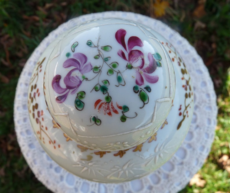 May include: A white porcelain lid with a floral design. The lid has a raised rim and is decorated with pink, purple, and green flowers. The lid is also decorated with gold accents.