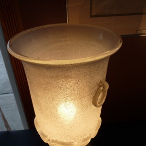 Puede incluir: Una pantalla de l&aacute;mpara de vidrio esmerilado con un asa decorativa. La pantalla est&aacute; iluminada desde el interior, proyectando un brillo c&aacute;lido.