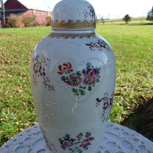 May include: A white porcelain vase with a lid, decorated with floral patterns in shades of red, pink, purple, and green. The vase has a heart-shaped design with flowers inside and a gold trim around the top.