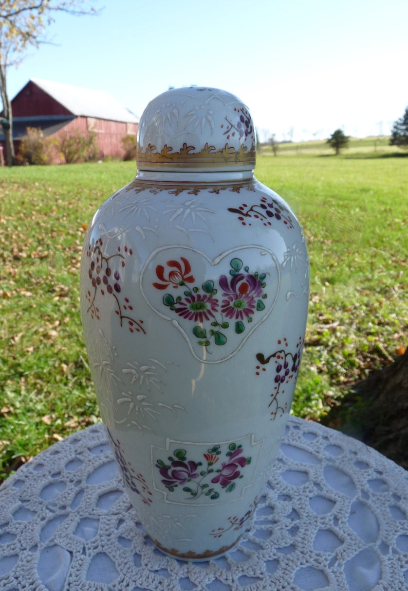 May include: A white porcelain vase with a lid, decorated with floral patterns in shades of red, pink, purple, and green. The vase has a heart-shaped design with flowers inside and a gold trim around the top.