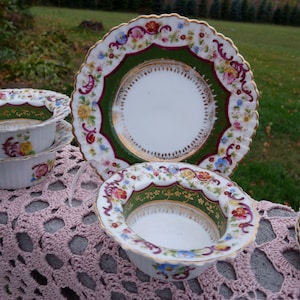 Puede incluir: Un juego de tres tazas de té y platillos de porcelana vintage con motivos florales. Las tazas y los platillos presentan intrincados bordes dorados y delicados diseños florales en tonos rosa, azul y verde. Los platillos tienen un borde festoneado y un diseño central con fondo verde.