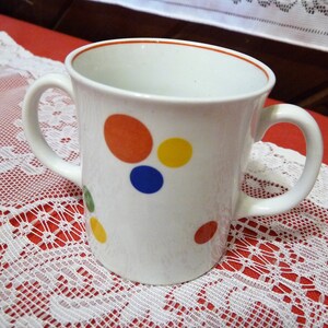 May include: White ceramic mug with two handles, featuring a red, yellow, blue, and green polka dot pattern.