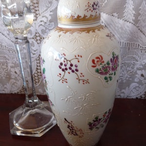 May include: A white porcelain vase with a gold rim and floral designs in shades of pink, purple, and red. The vase has a lid and is decorated with a delicate floral pattern.