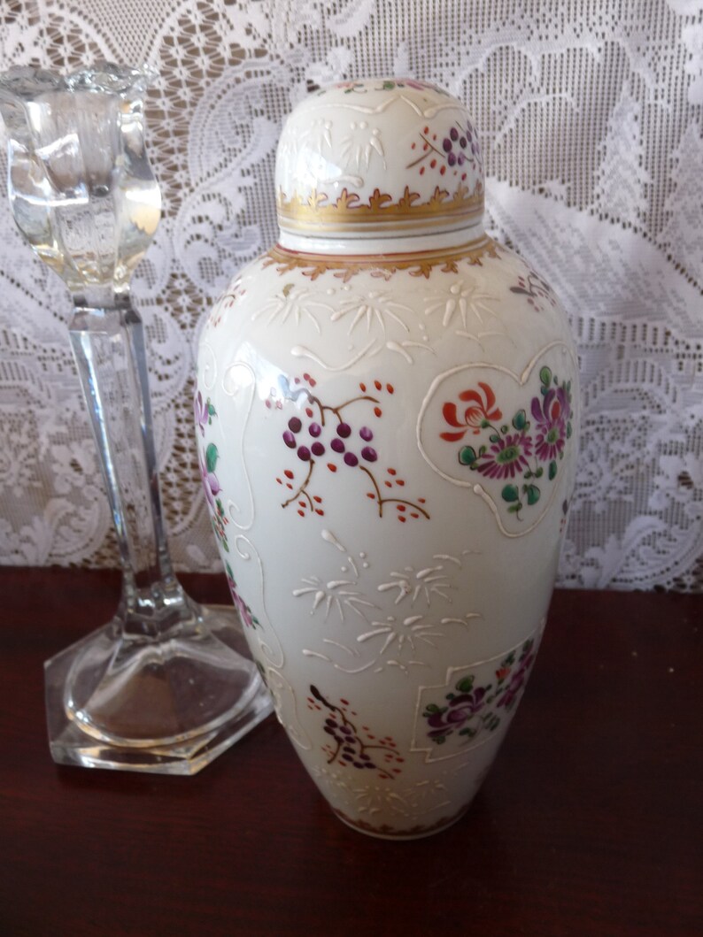 May include: A white porcelain vase with a gold rim and floral designs in shades of pink, purple, and red. The vase has a lid and is decorated with a delicate floral pattern.