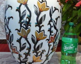 May include: Large white ceramic vase with a floral design in black, gold, and orange. The vase has a wide body and a narrow neck. A green Sprite bottle is next to the vase.