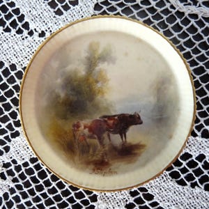 May include: A round, cream-colored decorative plate with a gold rim. The plate features a pastoral scene with two cows in a field, painted in muted tones. The artist's signature, "J. Slayton," is visible.