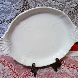 May include: A white ceramic serving tray with a decorative floral design. The tray has a scalloped edge and is displayed on a black stand.