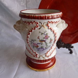 Op de afbeelding: Witte porseleinen vaas met rode en gouden accenten. De vaas heeft een decoratief ontwerp met een pauw en bloemen. De vaas heeft twee leeuwenkop handvatten.