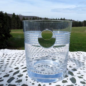 May include: A clear glass tumbler with a circular cut-out and horizontal ribbed detailing. The glass sits on a white, open-weave surface, with a backdrop of green grass, trees, and a blue sky.