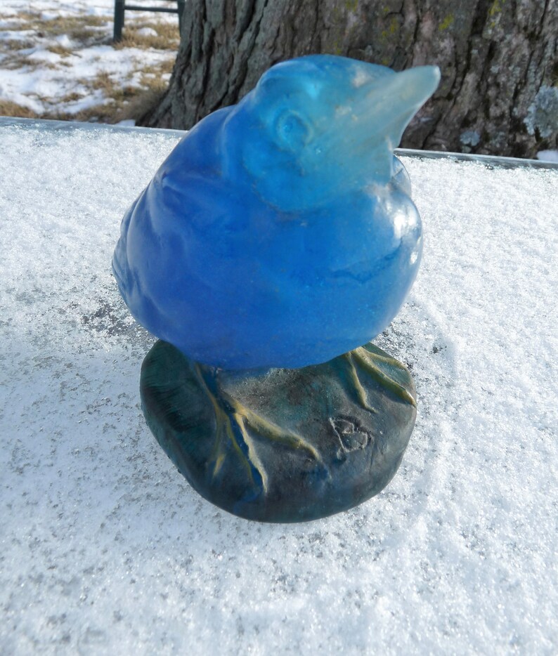 Pode incluir: Uma figurinha de p&aacute;ssaro de vidro azul com uma base detalhada. O p&aacute;ssaro est&aacute; empoleirado em uma base texturizada com uma pequena inscri&ccedil;&atilde;o indistinta.