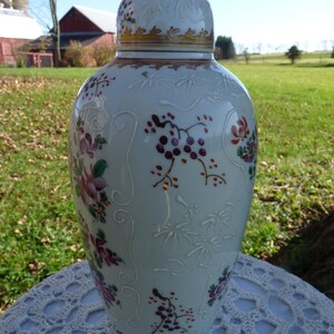 May include: A white porcelain vase with a gold rim and floral designs in shades of pink, purple, and green. The vase has a raised design and is decorated with a delicate floral pattern.
