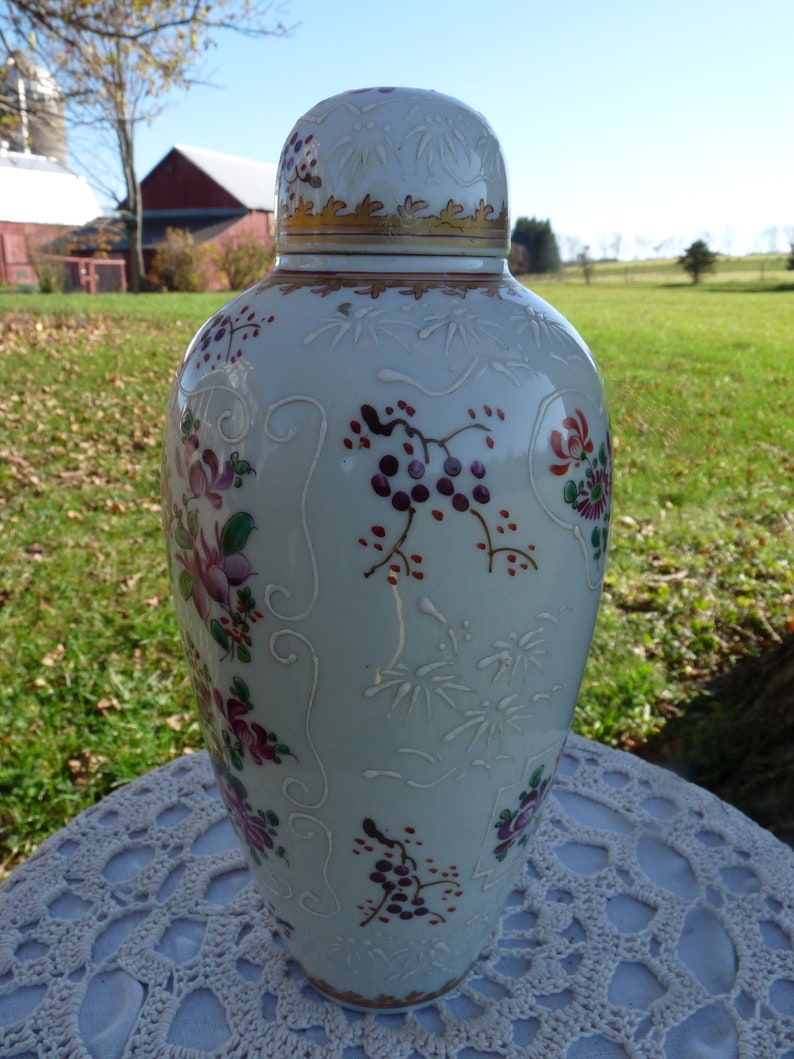 May include: A white porcelain vase with a gold rim and floral designs in shades of pink, purple, and green. The vase has a raised design and is decorated with a delicate floral pattern.