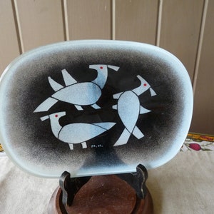 May include: A white oval glass dish with a black background and a design of three stylised birds in white with red eyes. The dish is on a wooden stand. The dish is signed "M.H."