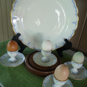 May include: Four white egg cups with blue and gold trim, each holding a different colored egg. The egg cups are on a wooden base and a larger white plate with blue and gold trim is in the background.
