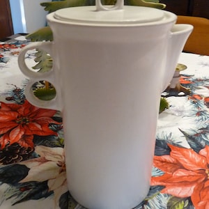 May include: White ceramic coffee pot with a lid and two handles. The pot is sitting on a table with a floral tablecloth.