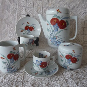 May include: A white porcelain tea set with a floral design. The set includes a teapot, creamer, sugar bowl, and two teacups and saucers. The flowers are red, blue, and green.
