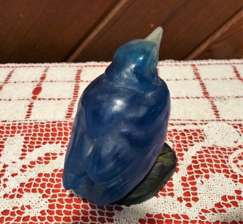 Pode incluir: Uma figurinha de p&aacute;ssaro de vidro azul com um acabamento liso e brilhante. O p&aacute;ssaro est&aacute; empoleirado em uma pequena base cinza escura.