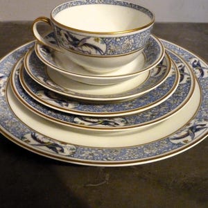 Puede incluir: Un juego de cuatro platos de porcelana blanca con diseños florales azules y dorados. Los platos están apilados uno encima del otro, con una taza de té y un platillo a juego en la parte superior.