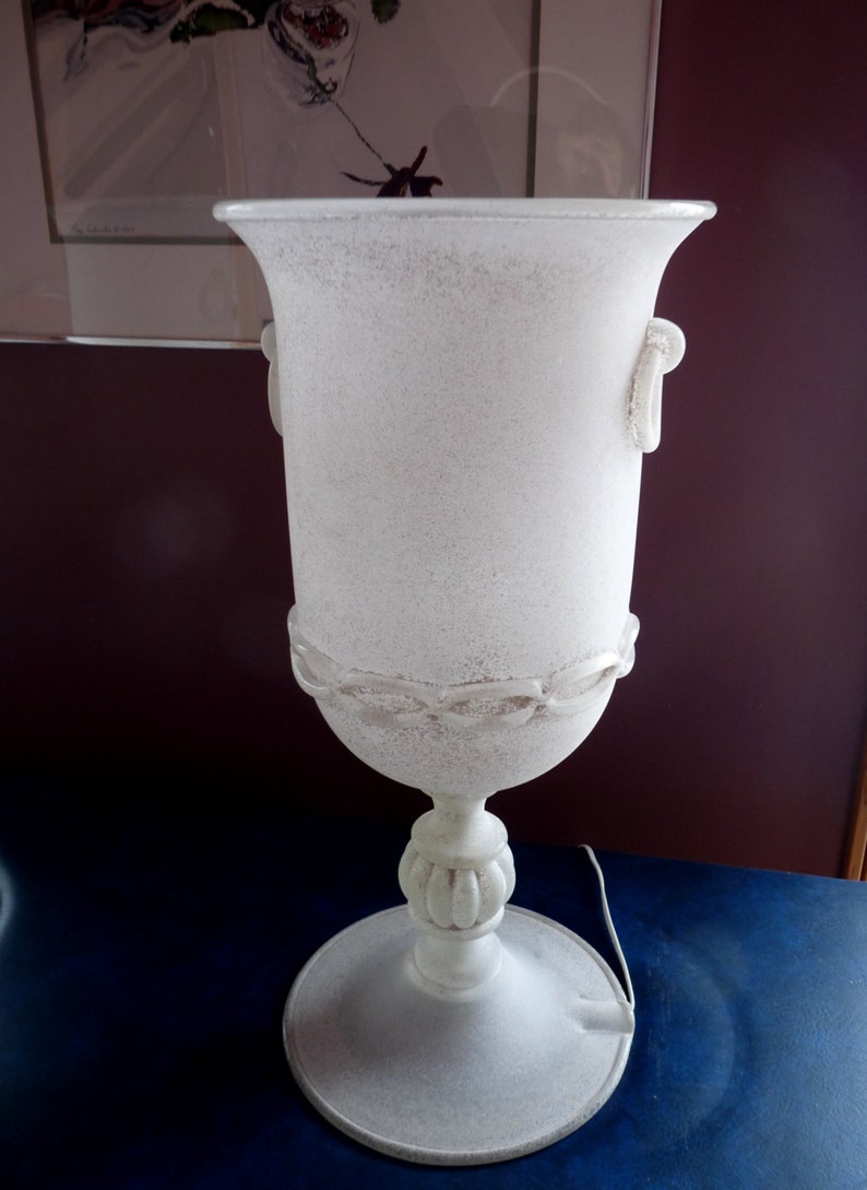Puede incluir: L&aacute;mpara de mesa de cer&aacute;mica blanca con un acabado texturizado y un dise&ntilde;o cl&aacute;sico. La l&aacute;mpara tiene una base ancha y un cuerpo alto y delgado con dos asas.