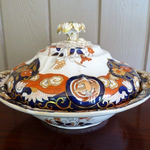 May include: A white porcelain serving dish with a lid, featuring a blue, orange, and gold floral pattern. The dish has a scalloped edge and a knob on the lid.