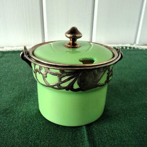 May include: A light green ceramic sugar bowl with a lid and a silver-colored filigree design around the rim. The lid has a small knob and a slot for a spoon. The bowl is sitting on a green surface.