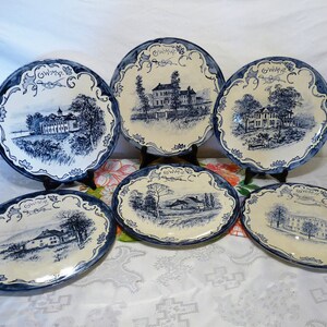 May include: Six blue and white ceramic plates with a decorative border and a black and white illustration of a building in the center. Each plate has the text "GW.M.P." in blue.