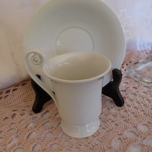 May include: White porcelain teacup and saucer set. The cup has a unique, curled handle and a classic design. The saucer is plain white.