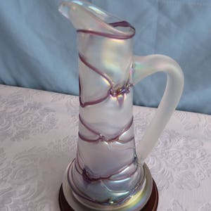May include: A clear glass pitcher with a frosted finish and iridescent purple and blue accents. The pitcher has a wide mouth and a curved handle. It sits on a wooden base.