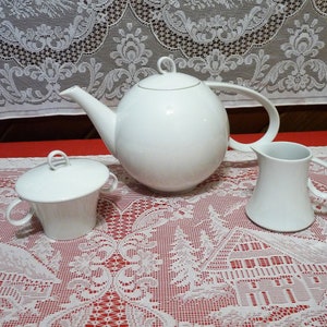 May include: A white teapot, creamer and sugar bowl with lids. The teapot has a round body and a curved spout. The creamer and sugar bowl have simple, rounded shapes. All three pieces are made of white porcelain.