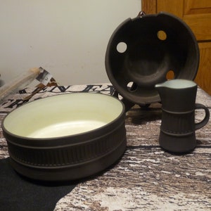 以下が含まれることがあります： 黒色のセラミック製のキッチン用品3点セット。薄いベージュ色の内側の丸いボウル、薄い水色の内側の小さなピッチャー、蓋付きで上面に4つの穴が開いた鍋です。