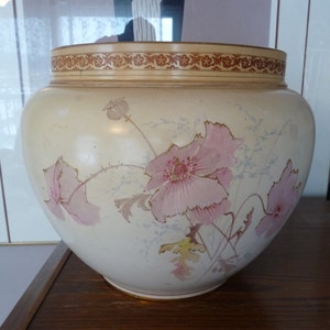 Puede incluir: Un macetero grande de cerámica de color crema con un diseño floral. El macetero tiene un borde marrón y dorado y presenta flores rosas con hojas verdes.