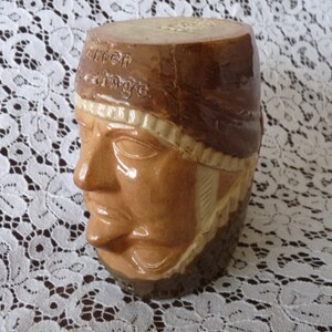 May include: A brown ceramic mug shaped like a person's head with a brown and white hat. The hat has the words "Allen Carriage" written on it.