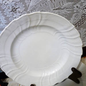 May include: A white, textured ceramic plate with a scalloped edge and a decorative wave pattern. The plate is round and appears to be part of a dinnerware set. The plate is on a dark brown stand.