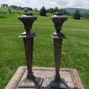 May include: Two bronze-colored, ornate candle holders with square bases and fluted columns. The tops have a decorative, urn-like design. They are placed on a woven surface, with a green field and blue sky in the background.