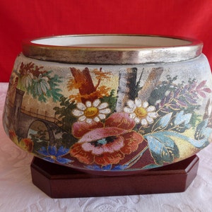 May include: A decorative ceramic bowl with a landscape design featuring a bridge, trees, and flowers. The bowl has a silver rim and sits on a dark brown wooden base. The floral design includes daisies and other colorful blooms.