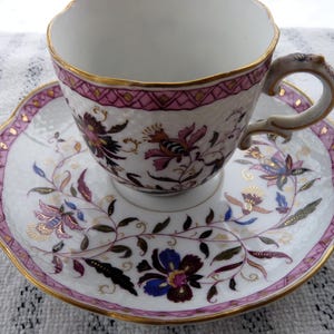 May include: A white teacup and saucer set with a floral design. The cup and saucer have a pink and gold trim. The floral design includes purple, blue, and gold accents. The cup has a decorative handle.