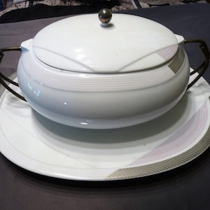 Op de afbeelding: Witte porseleinen serveerschaal met een deksel en twee metalen handvatten. De schaal heeft een zilveren en roze rand. De schaal staat op een bijpassend bord.