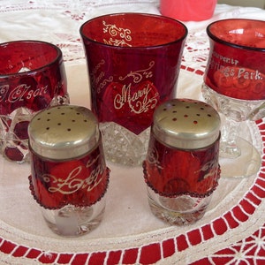 Puede incluir: Una colección de cristalería de color rojo rubí, que incluye una taza, dos saleros y dos vasos. La cristalería presenta letras y diseños grabados decorativos. Los saleros tienen tapas plateadas y adornos de cuentas.