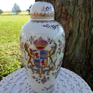 May include: A white porcelain vase with a lid, decorated with a coat of arms featuring a lion and a unicorn, surrounded by floral motifs in shades of pink, red, and gold.