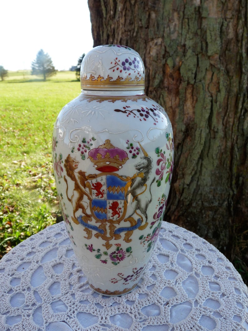 May include: A white porcelain vase with a lid, decorated with a coat of arms featuring a lion and a unicorn, surrounded by floral motifs in shades of pink, red, and gold.