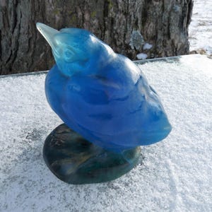Pode incluir: Uma escultura de vidro azul de um p&aacute;ssaro com a cabe&ccedil;a inclinada para baixo. O p&aacute;ssaro est&aacute; empoleirado em uma base verde.