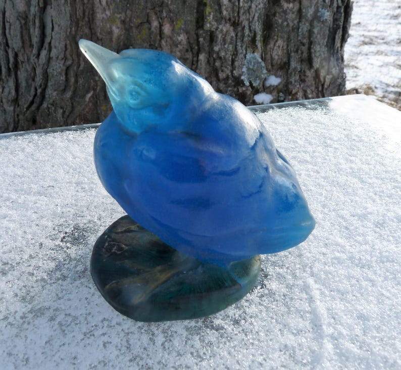 Pode incluir: Uma escultura de vidro azul de um p&aacute;ssaro com a cabe&ccedil;a inclinada para baixo. O p&aacute;ssaro est&aacute; empoleirado em uma base verde.