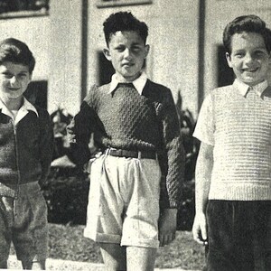 Op de afbeelding: Zwart-witfoto van drie jongens in vintage kleding. Ze dragen truien, overhemden met kraag en shorts. De jongens staan voor een gebouw en poseren voor de camera.