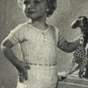 Op de afbeelding: Een jong kind in een lichtgekleurde, gebreide romper met korte mouwen en een korte broek. De romper heeft een knoopsluiting en een textuurpatroon. Het kind houdt een knuffel vast.