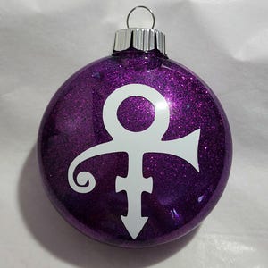 May include: A purple glitter Christmas ornament with a white symbol. The ornament has a silver metal cap and a loop for hanging. The symbol is a stylized combination of male and female symbols, often associated with the musician Prince.