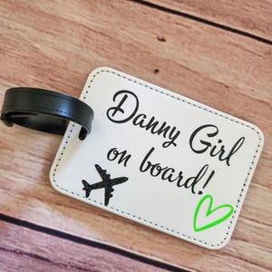 May include: White luggage tag with black stitching and a black strap. The tag has black text that says "Danny Girl on board!" with a black airplane icon and a green heart.