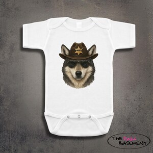 May include: White baby bodysuit featuring a wolf wearing a brown sheriff hat and sunglasses. The wolf has a detailed fur design. The hat has a gold star badge. The bodysuit has snap closures at the bottom.